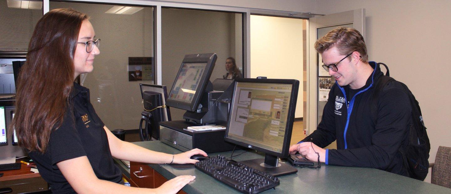 年轻女子站在桌子后面，手里拿着电脑, 在柜台的另一边帮一个年轻人